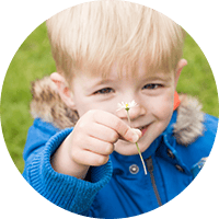 Young boy smiling outdoors holding a small flower, enjoying nature, representing childhood happiness and outdoor learning at Thrive Childcare.