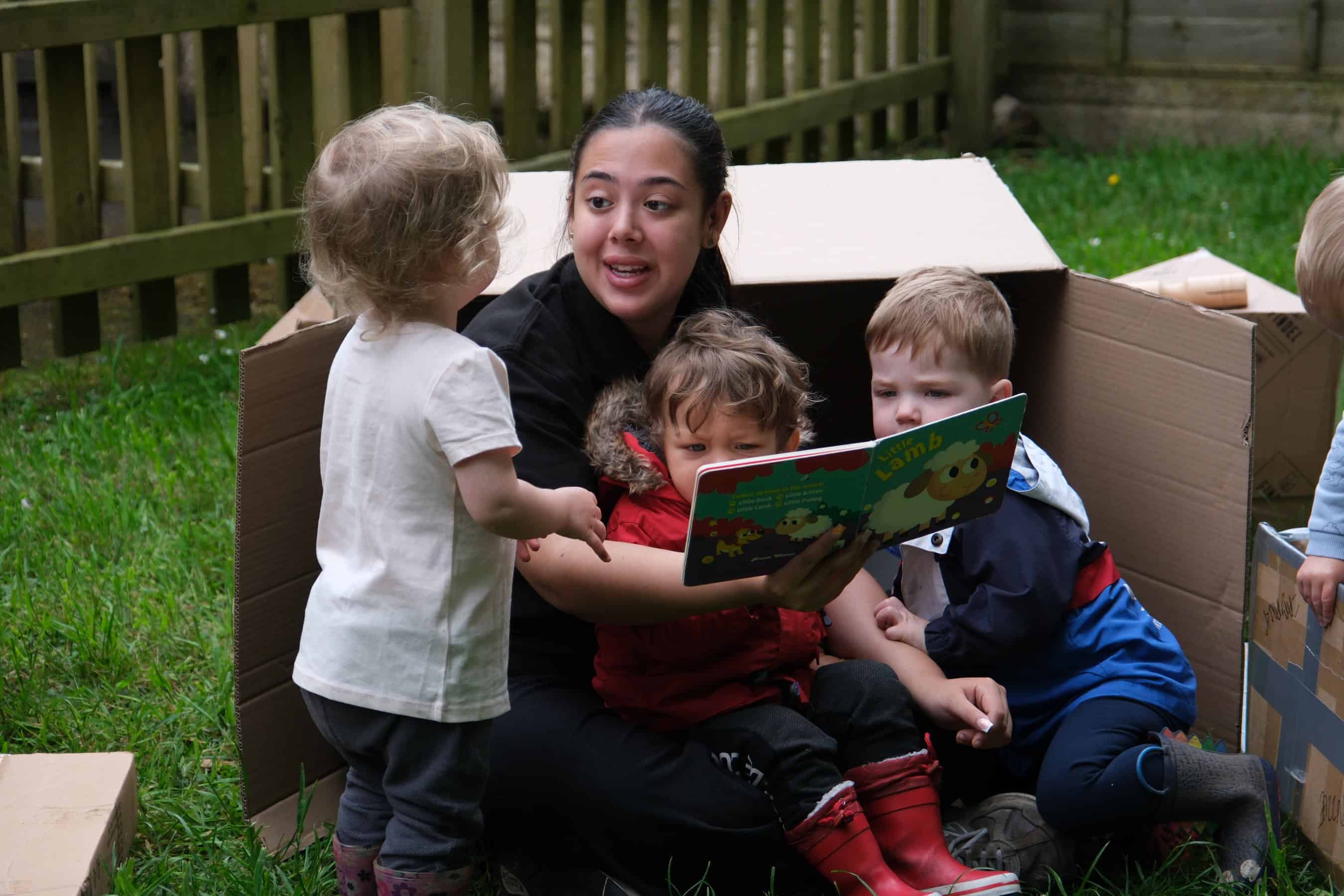 Children's outdoor playtime with caregiver reading a story in a cardboard playhouse, promoting early childhood education and social development at Thrive Childcare.
