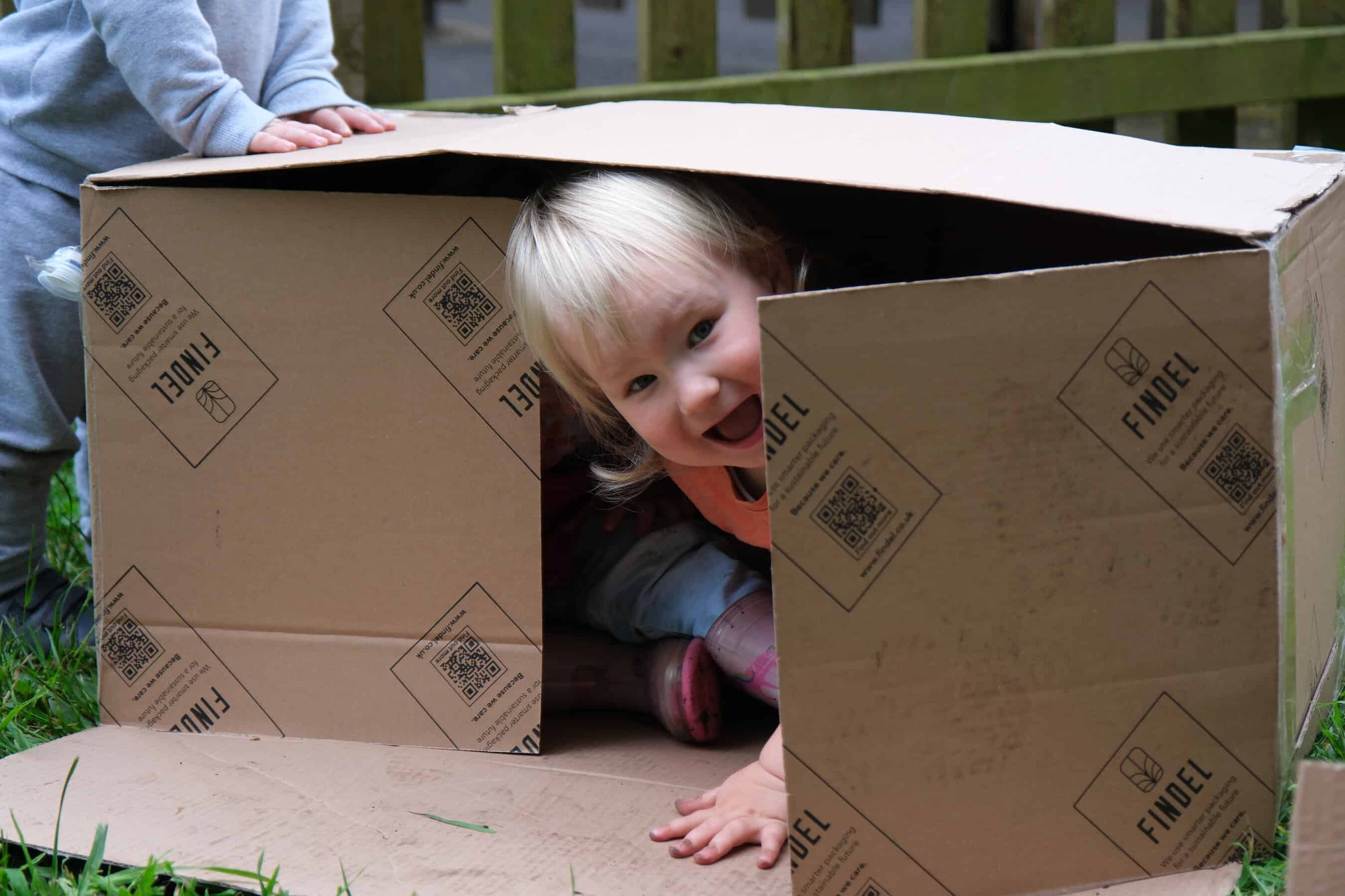 A young child playing or exploring inside a large cardboard box outdoors, demonstrating creativity and active learning at Thrive Childcare.