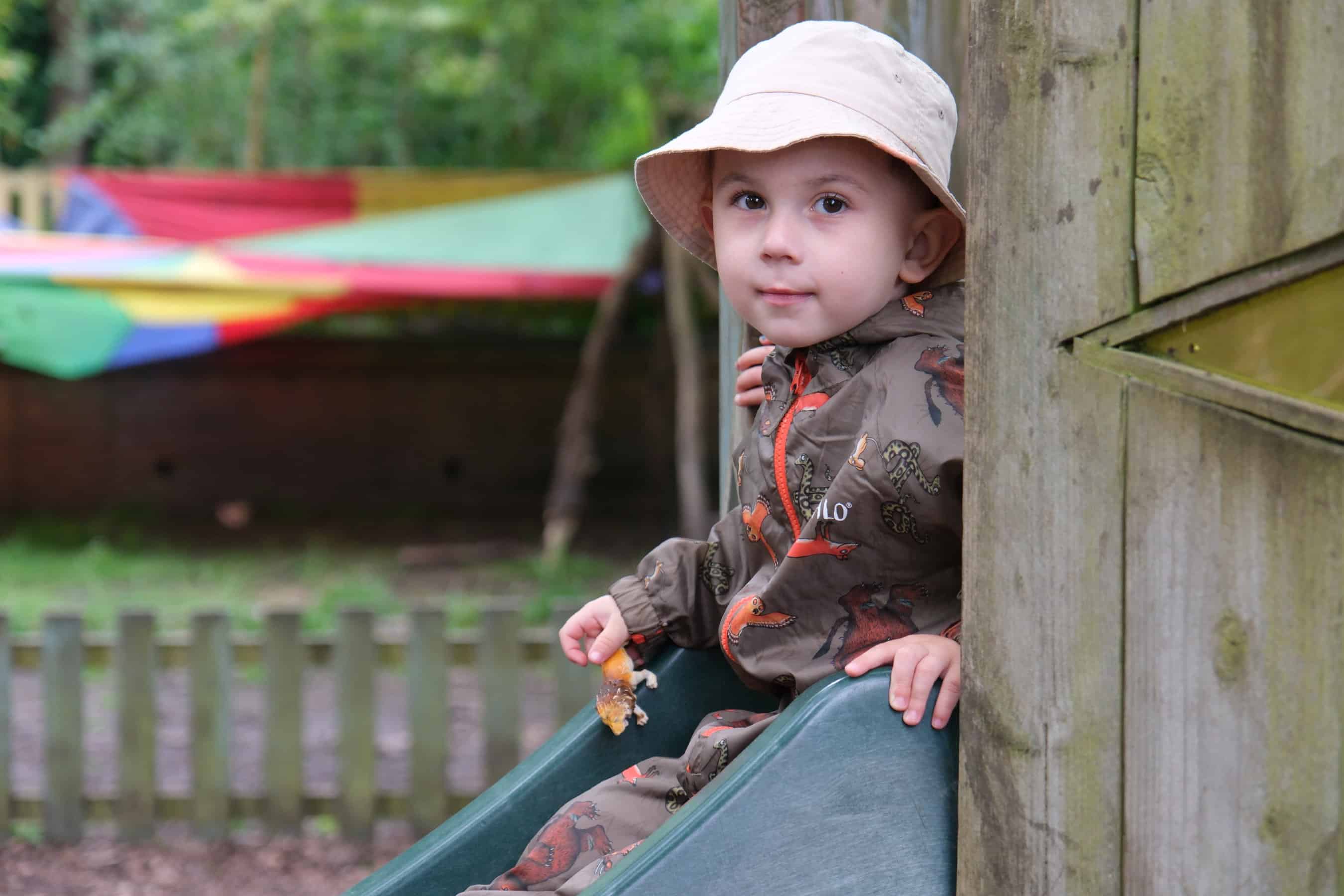 Young child wearing a sun hat and raincoat sitting on a slide at Thrive Childcare outdoor play area in nature setting.