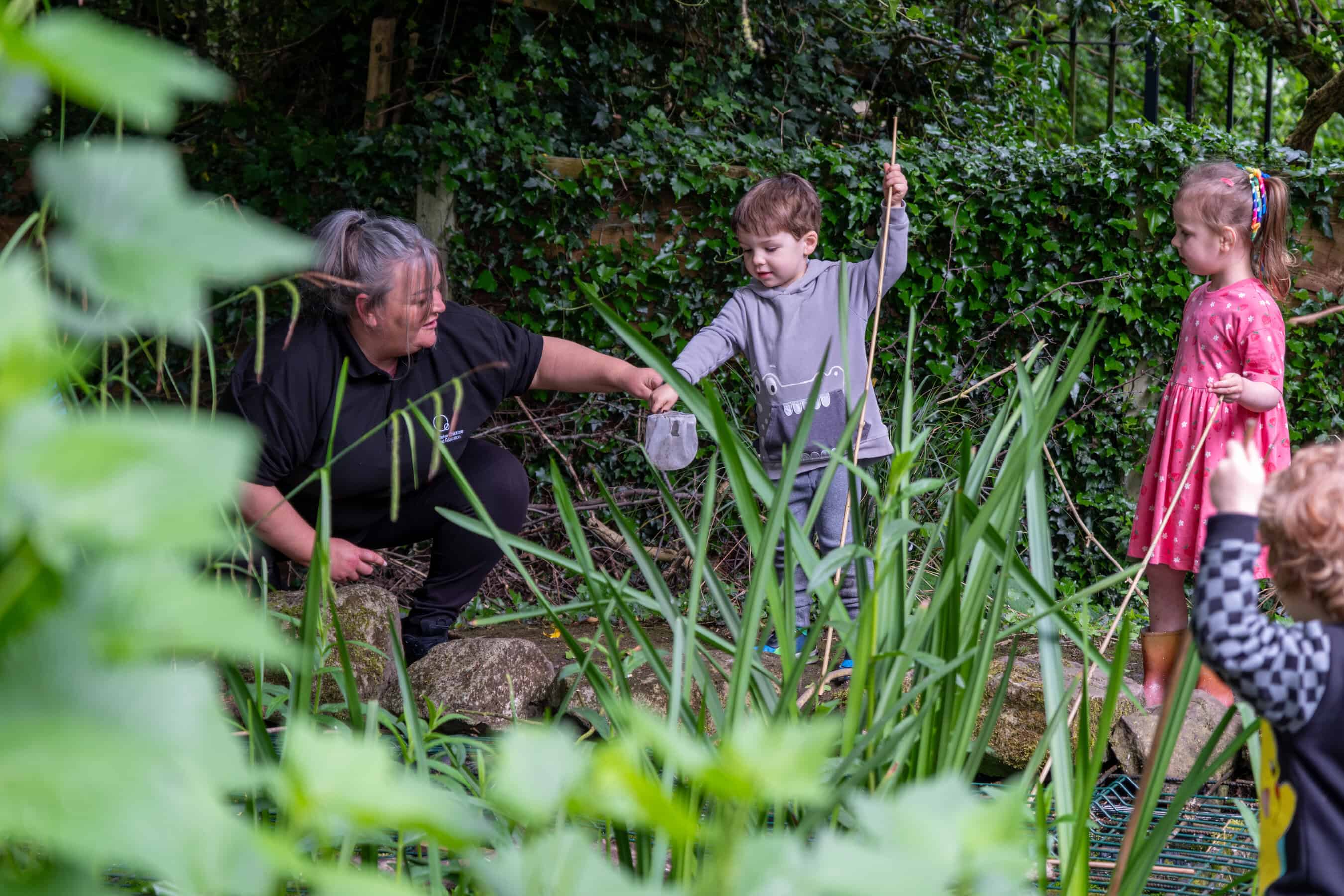 Children engaging in outdoor nature activities at Thrive Childcare, exploring plants and wildlife in a lush, green environment with caring staff.