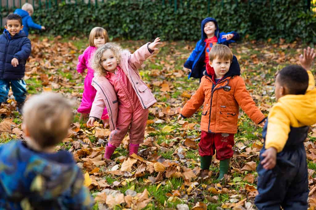 Brightly dressed children playing outdoors on autumn leaves at Thrive Childcare, promoting outdoor activities, early childhood development, and safe, engaging childcare environments.