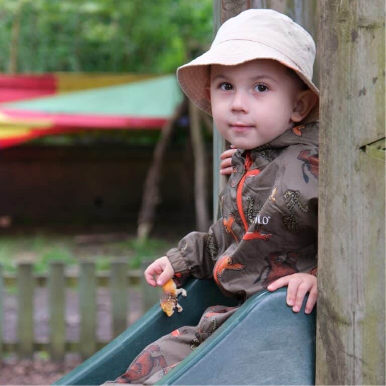 Little child in colourful outdoor playground at Thrive Childcare, dressed in a dinosaur print jacket and a sun hat, enjoying playtime on a slide in a safe and nurturing environment.