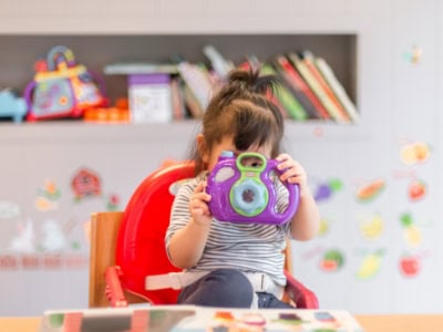 Bright-eyed young girl holding a colourful camera toy while sitting at a table in a vibrant classroom at Thrive Childcare, fostering creativity and early development.