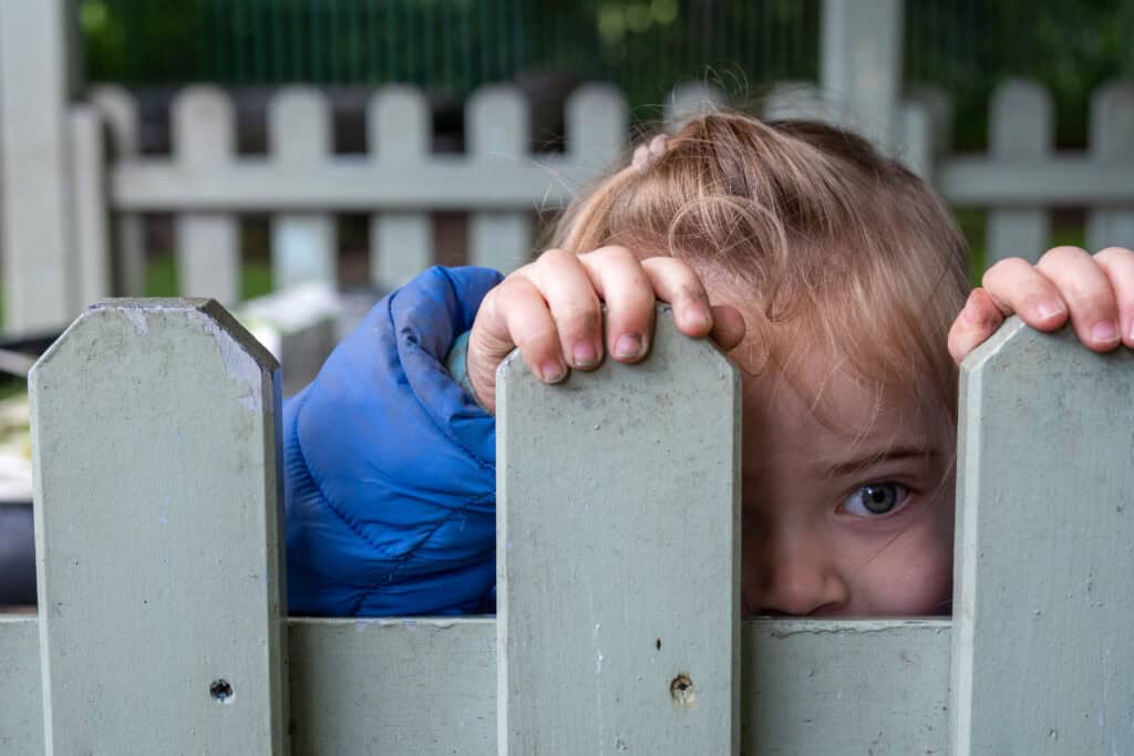 1. Young girl peeking through a wooden fence at Thrive Childcare, outdoor play area, children's safety and supervision, early childhood education, happy kids, childcare services.