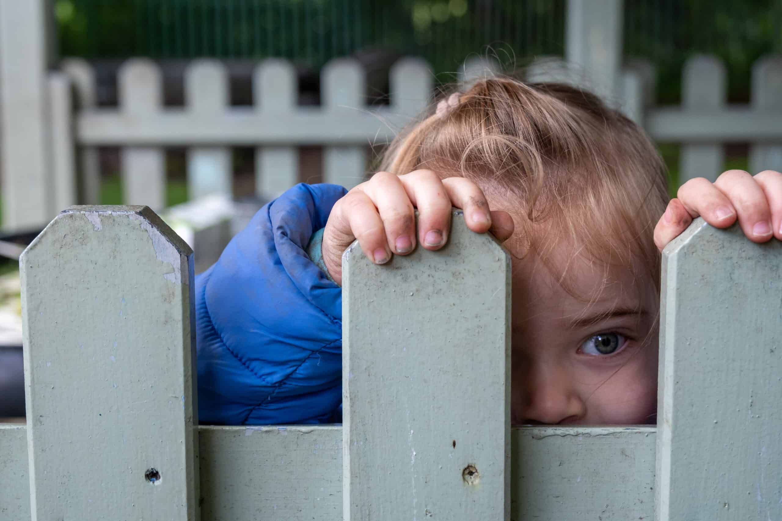 1. Young girl peeking through a wooden fence at Thrive Childcare, outdoor play area, children's safety and supervision, early childhood education, happy kids, childcare services.