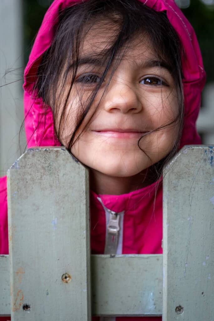 Bright smiling girl in pink jacket peeks over wooden fence, joyful child enjoying outdoor play at Thrive Childcare, colourful and caring environment for early childhood development and fun learning activities.