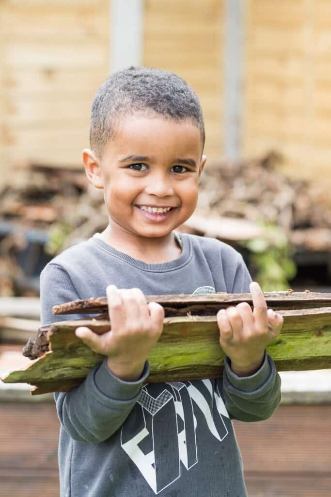 Bright smiling boy holding a large piece of wood outdoors, promoting safe childcare and outdoor play at Thrive Childcare for happy, confident children.