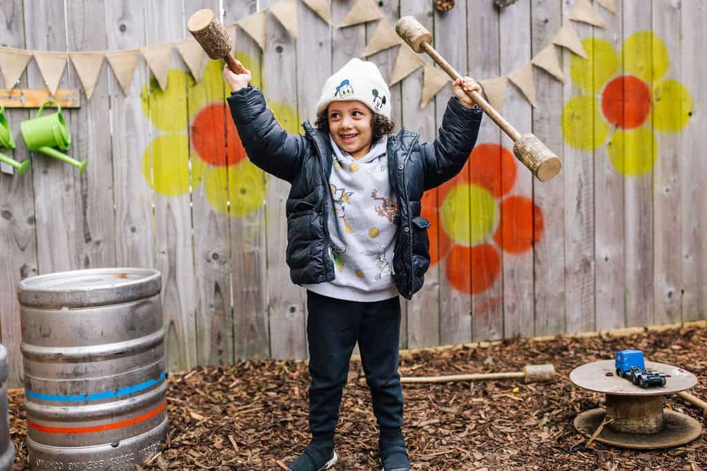 Bright-eyed child playing outdoors in a colourful nursery environment, enjoying fun with wooden hammers, highlighting Thrive Childcare’s engaging early childhood education and outdoor play activities.