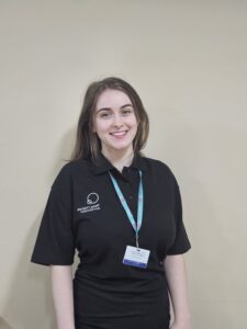 A smiling young woman wearing a Thrive Childcare uniform and name badge, standing against a beige wall, representing professional childcare staff dedicated to early childhood education and child development.