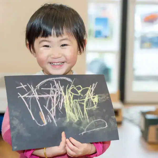 Smiling young child holding colourful chalk drawing on black paper in childcare setting, demonstrating early childhood education and creative play at Thrive Childcare.