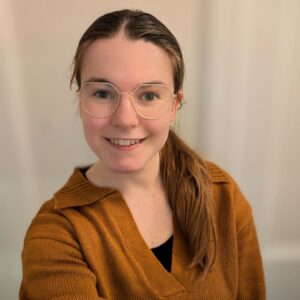 Warm smile of a female childcare professional with glasses, wearing a mustard cardigan, in a well-lit environment. Focused on supporting child development at Thrive Childcare.