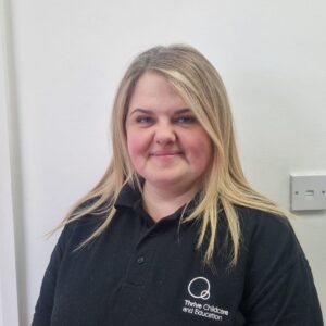 High-quality childcare professional wearing Thrive Childcare uniform, standing indoors; dedicated to supporting early childhood education and development. Suitable for childcare, preschool, and education settings.