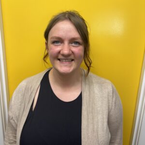 A friendly childcare worker at Thrive Childcare smiling in front of a vibrant yellow background, showcasing caring staff dedicated to early childhood development and quality childcare services.