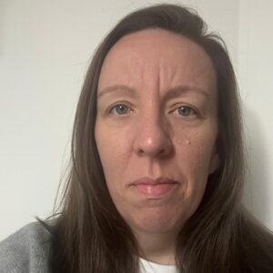 A woman with long brown hair and blue eyes, looking directly at the camera, standing indoors against a plain white wall, representing caring and professional childcare environment.