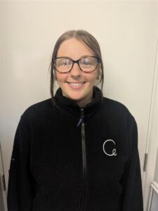 1. A smiling female childcare professional wearing glasses and a black Thrive Childcare uniform, standing against a plain cream-colored wall.