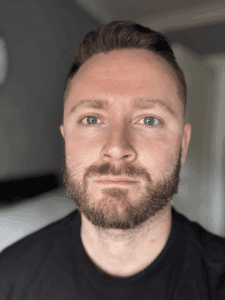 Bright adult man with short dark hair and beard, wearing a black shirt, in modern indoor setting—adventure, self-care, or professional profile image for Thrive Childcare.