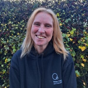 Bright smiling childcare professional wearing a Thrive Childcare hoodie, standing outdoors amidst autumnal foliage, promoting quality early childhood education and caring childcare services.