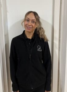 Caring childcare educator wearing Thrive Childcare branded jacket, smiling confidently in a bright indoor setting, exemplifying professional and nurturing childcare environment.