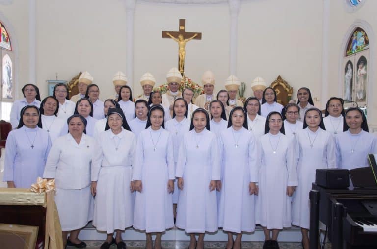 Nuns in white habits gathered in a church with a crucifix and stained glass windows, celebrating faith and community at Wildflower Home.