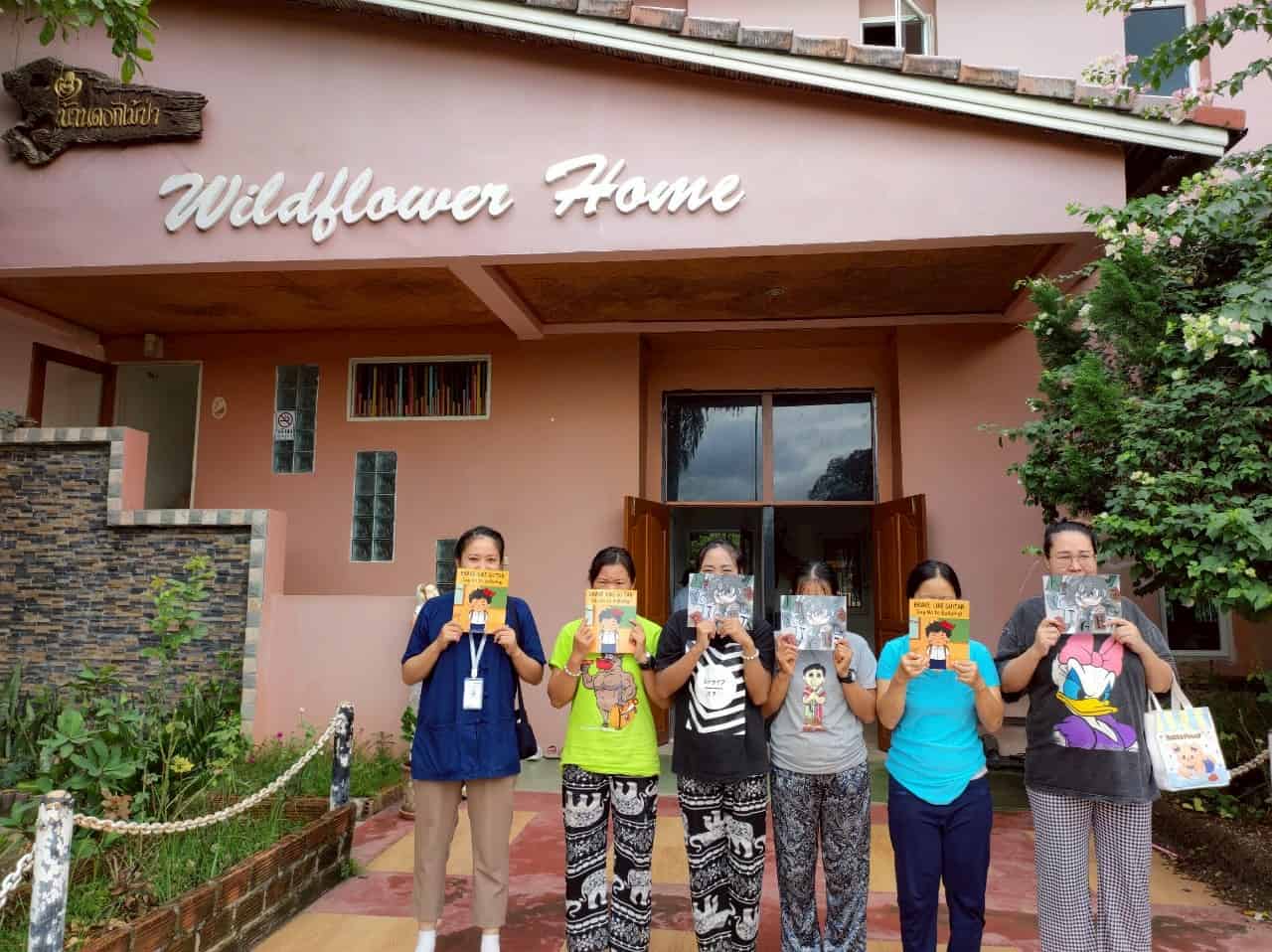 People outdoor group of women standing in front of pink building reading colorful illustrated books about guitar and cartoon characters, casual clothing, greenery, and welcoming atmosphere at Wildflower Home, inspiring community engagement and literacy.