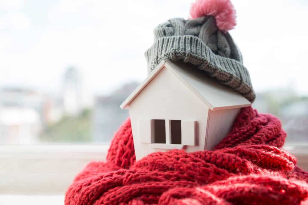 A small model house is wrapped in a red knitted scarf and topped with a gray beanie, symbolizing warmth and coziness.