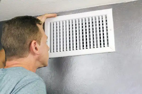 Person inspecting a white air vent on a gray wall.