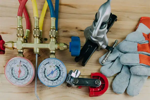HVAC tools on a wooden surface: manifold gauge set with red, blue, and yellow hoses, pipe bender, copper tubing cutter, and gray work gloves.