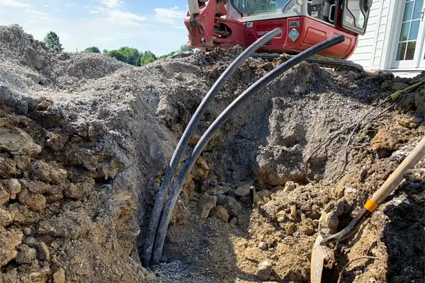 Excavated site with electrical cables emerging from the ground near a building, a red excavator, dirt mounds, and a shovel visible.