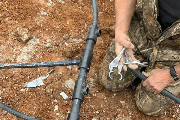 Person in camouflage suit repairs black plastic pipes on reddish soil, using a hand tool to connect them.