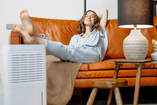A person relaxes on a tan sofa with feet up, eyes closed, and arms behind their head. A dehumidifier is in the foreground, and a lamp is on a side table.
