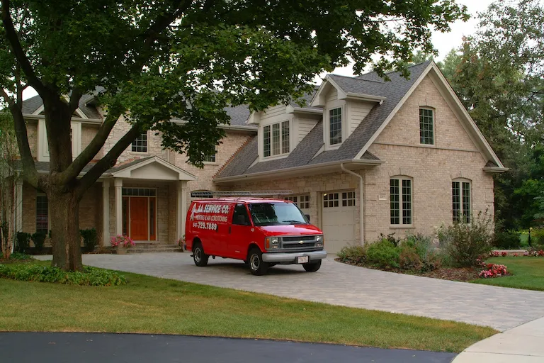 A red plumbing service van is parked in the driveway of a large, two-story brick house, possibly for an indoor air quality installation. Trees and a well-maintained lawn surround the property, enhancing its charm.