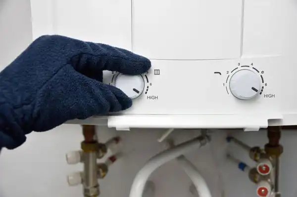 A gloved hand adjusts the temperature dials of a water heater, indicating "HIGH" settings, with visible pipes and valves below.