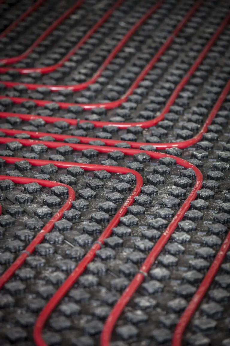 Red pipes are installed in a zigzag pattern within a black grid on a floor, likely part of an underfloor heating system.