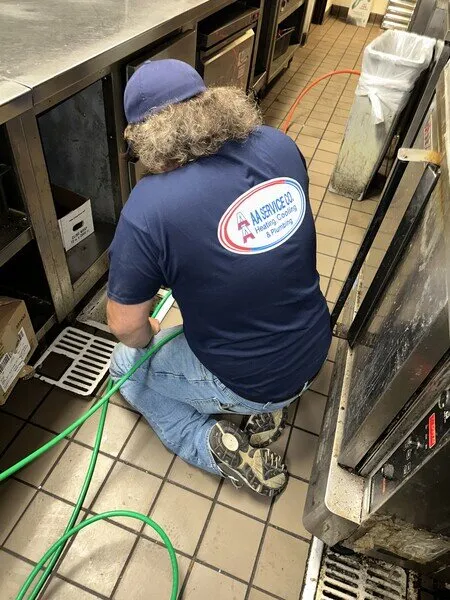 Person in a blue shirt and cap is kneeling on a tiled floor, working with a green hose behind kitchen equipment.