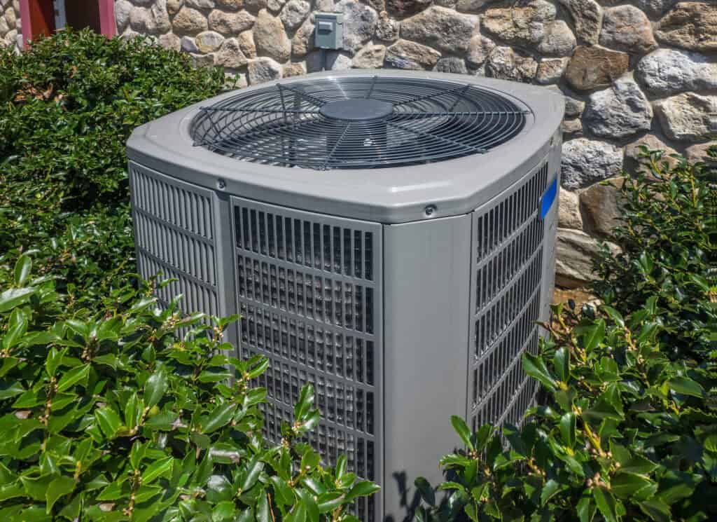 The outdoor air conditioning unit, provided by Cooling Services, nestles amid lush green bushes near a stone wall of the building.