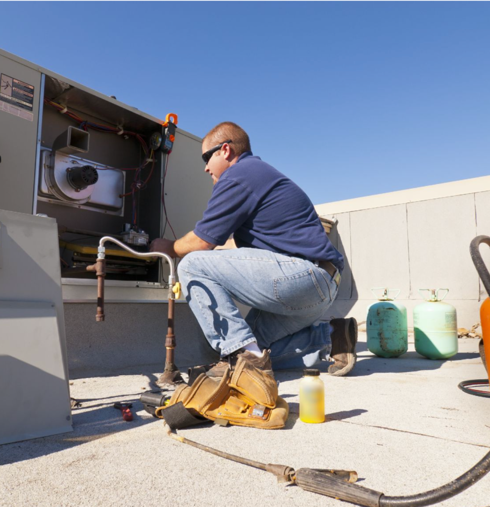 A technician in a blue shirt and jeans expertly works on an HVAC unit, delivering top-notch cooling services under a clear sky. Nearby, two gas cylinders and a tool bag rest on the rooftop.