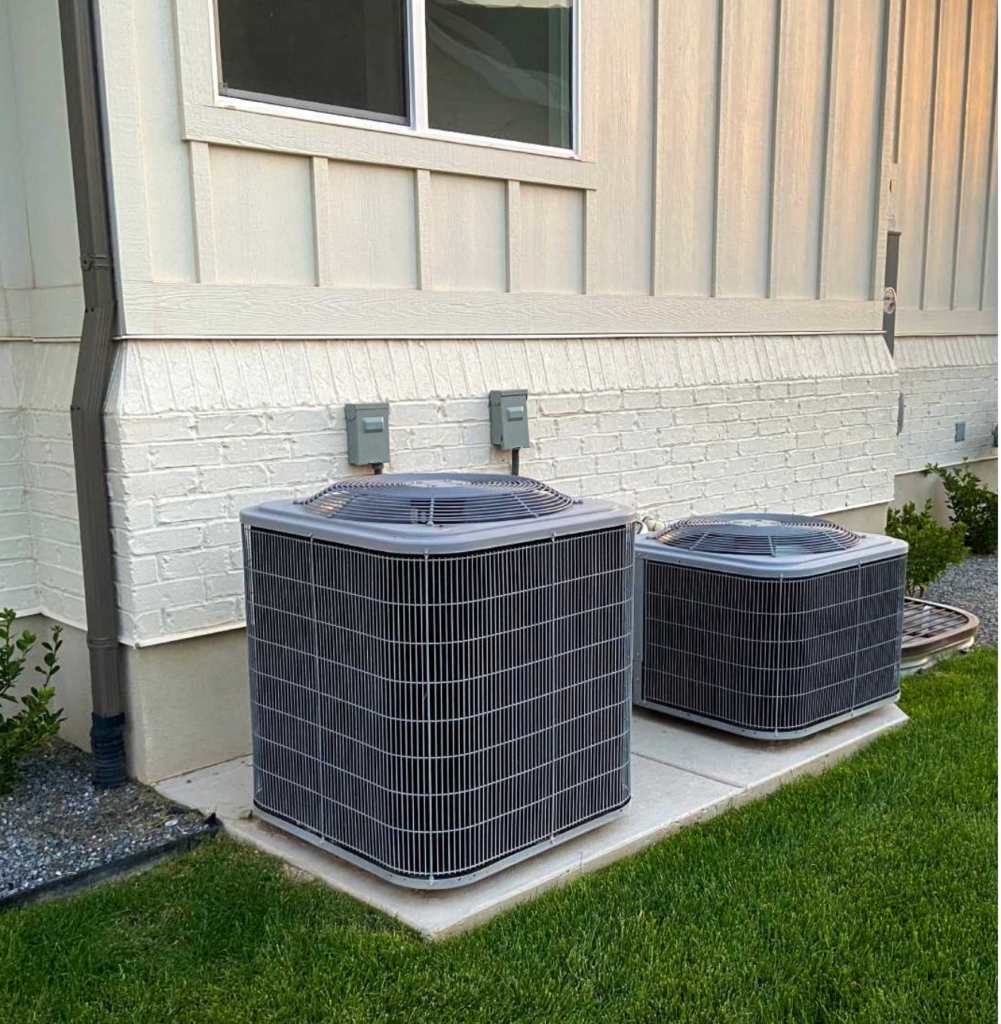 Two outdoor air conditioning units, part of our expert cooling services, rest on a concrete slab near a house with beige siding and brick. Green grass surrounds the area, offering a serene backdrop.
