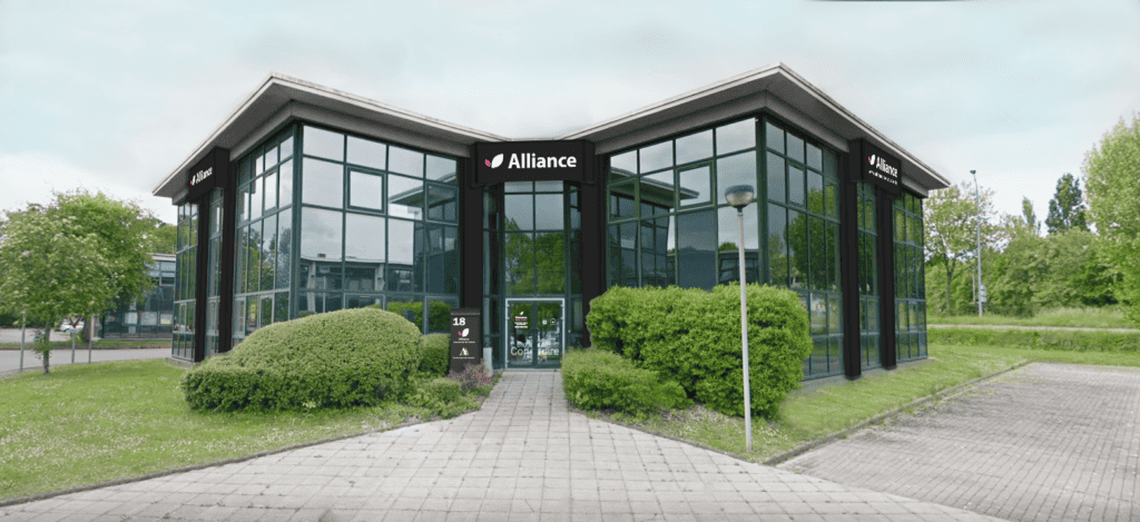 agence-nantes Façade d’un bâtiment professionnel moderne entièrement vitré, abritant une agence Alliance, avec un aménagement paysager verdoyant à l’avant et un parking adjacent.