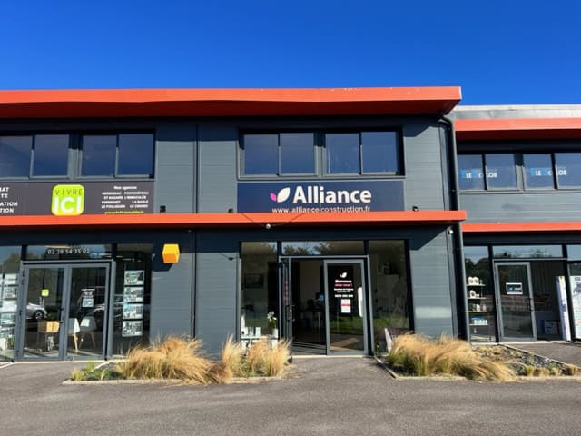 agence-pontchateau Façade moderne d’une agence Alliance Construction avec un bardage gris anthracite, une enseigne visible, des vitrines lumineuses et des aménagements paysagers à l’avant, située dans un bâtiment commercial.