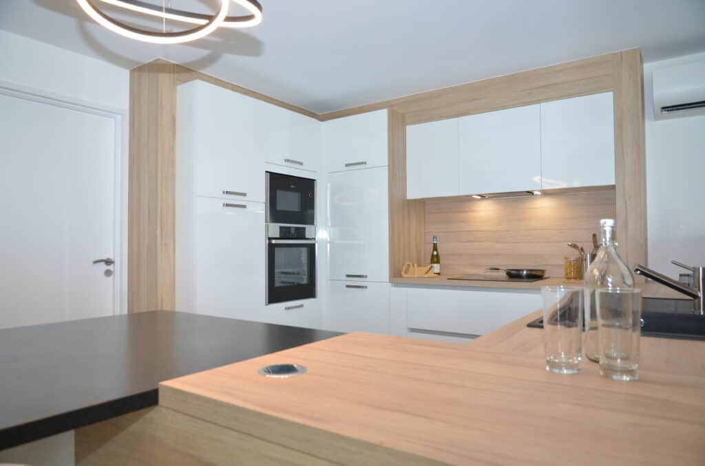 Cuisine contemporaine avec des façades blanches brillantes, des éléments en bois clair, un plan de travail en contraste noir, et un éclairage intégré, offrant un design épuré et fonctionnel.
