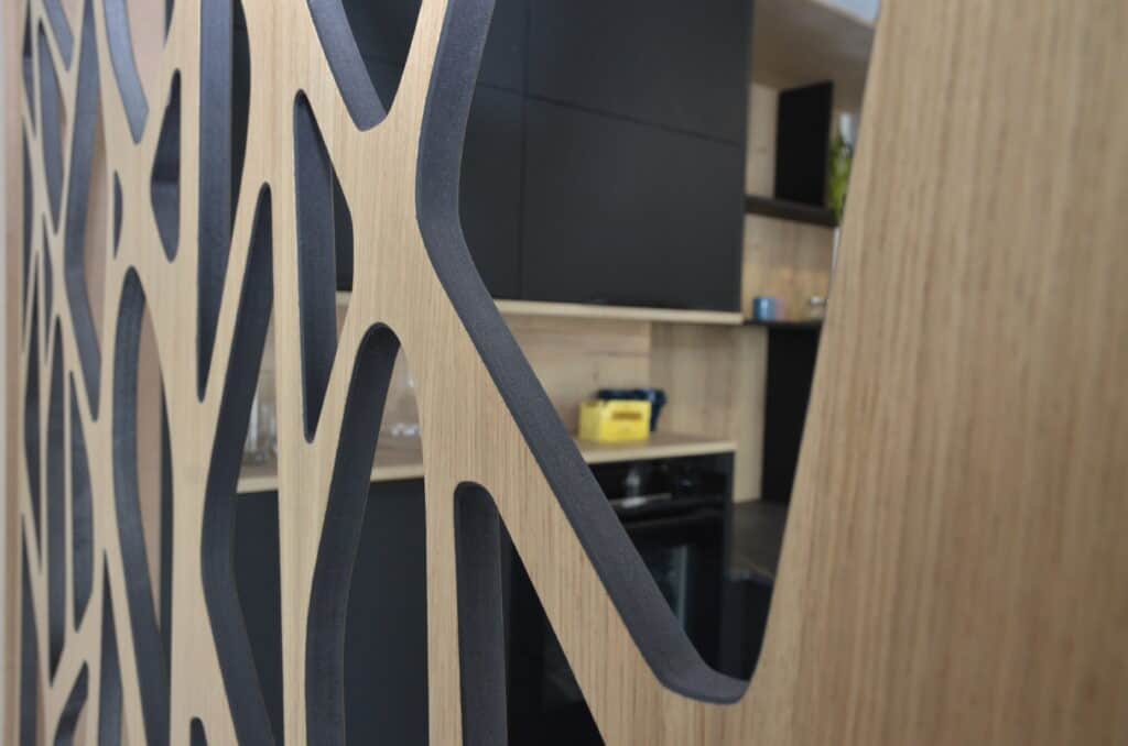 Gros plan sur une cloison décorative en bois ajouré avec des motifs organiques, intégrée dans une cuisine moderne avec des rangements en noir mat et un fond en bois clair.