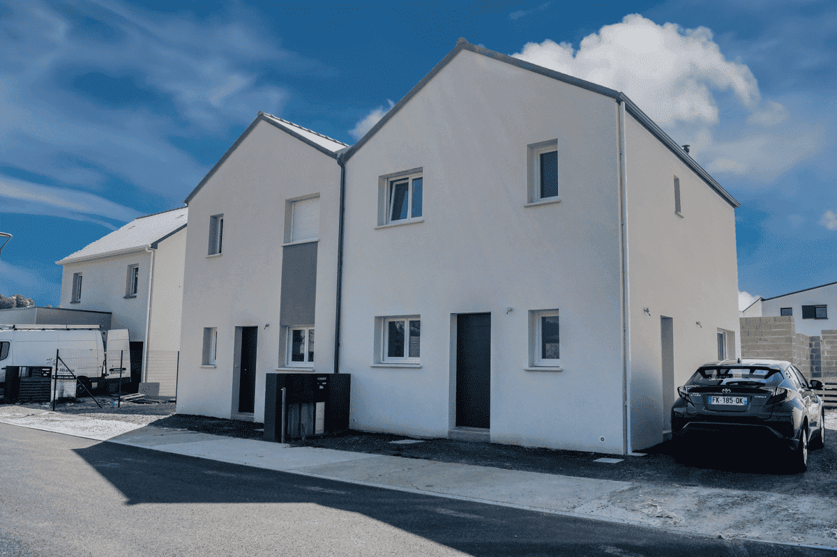 Maison mitoyenne moderne avec façade blanche, toit à deux versants et voiture stationnée devant, sous un ciel bleu lumineux.