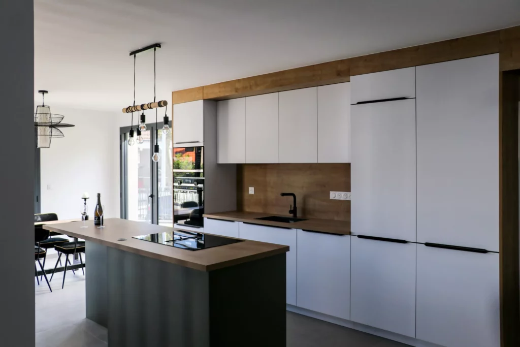 Cuisine contemporaine avec îlot central, suspensions design, mobilier blanc et bois, et électroménager intégré.