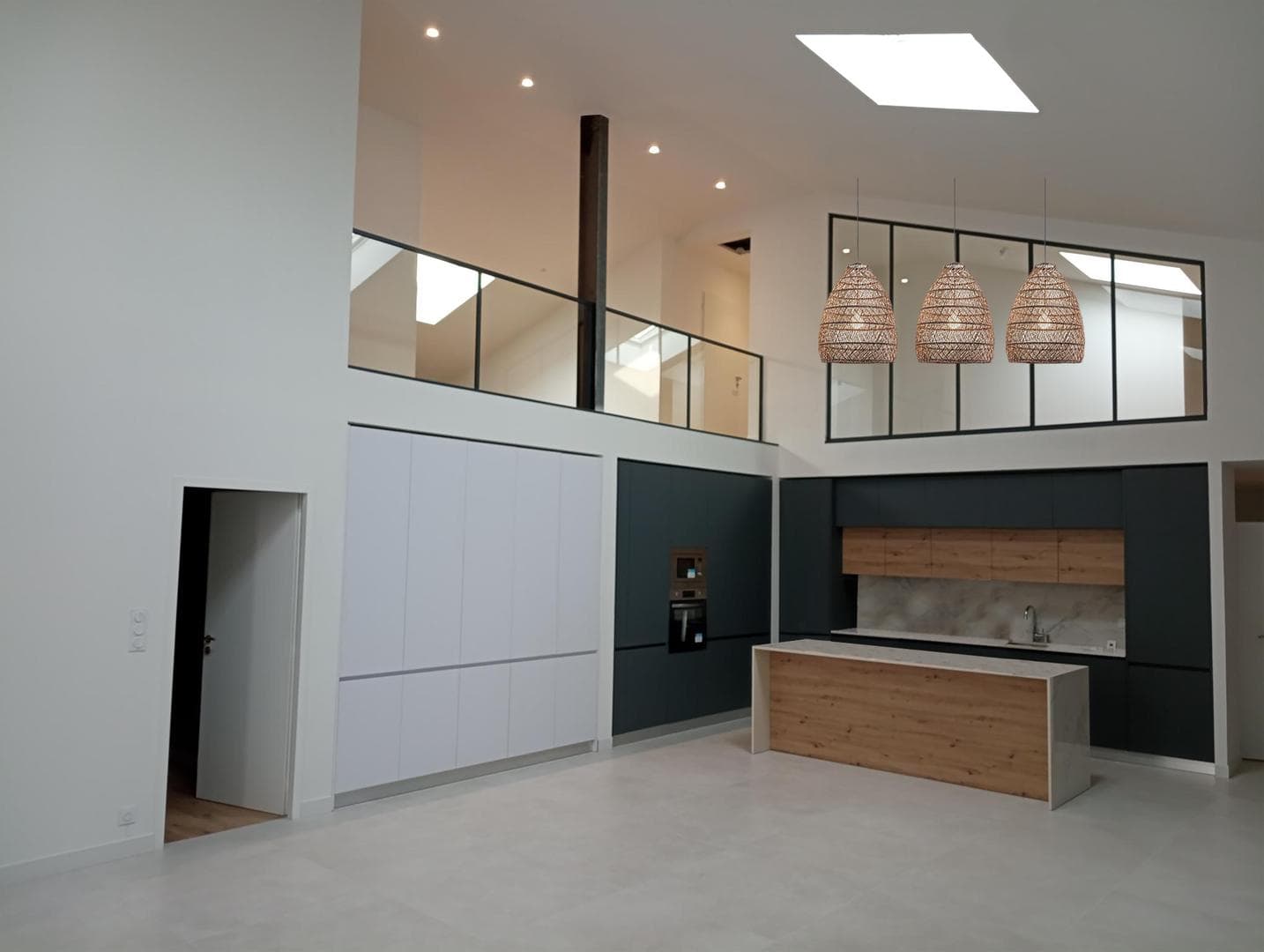 Cuisine moderne réalisée dans un espace lumineux avec un plafond haut. L'aménagement comprend des meubles de cuisine intégrés, une crédence en marbre et un îlot central en bois avec des éléments noirs. Des luminaires suspendus en osier ajoutent une touche chaleureuse. La grande baie vitrée et le puits de lumière apportent de la lumière naturelle dans cette pièce rénovée.