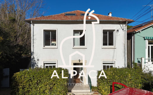 Maison blanche à deux étages, toit rouge, entourée de verdure, avec logo Alpaca Immobilier.
