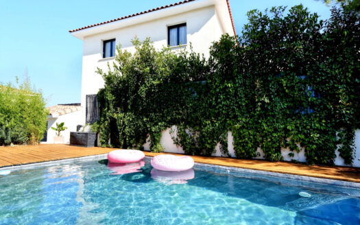 Maison blanche à toit rouge près de piscine avec bouées roses, entourée de verdure.