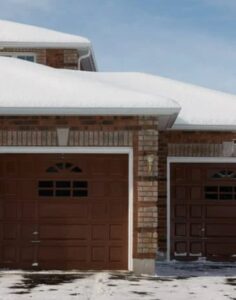 Snow-covered roof of a brick house with two brown garage doors, surrounded by a snowy driveway.