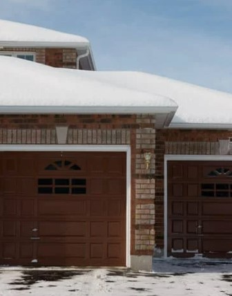Snow-covered roof of a brick house with two brown garage doors, surrounded by a snowy driveway.