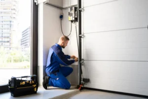 A technician in blue overalls kneels to repair a garage door system, with tools in a black bag nearby.